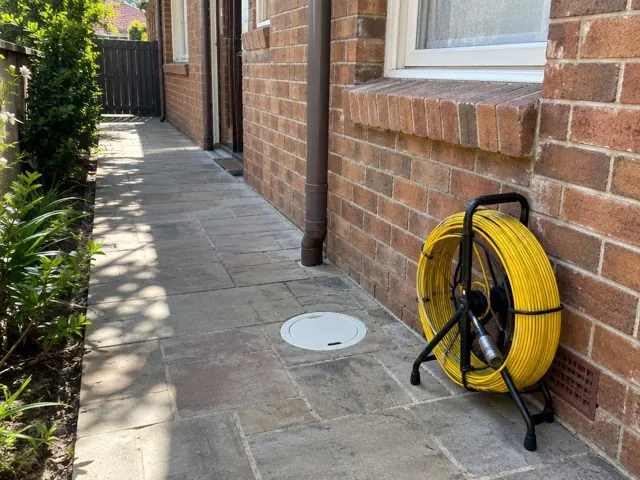 Capped inspection opening beside a yellow drain camera reel on a paved brick side path in Kensington