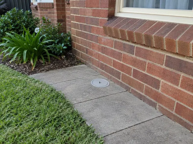 Capped access point set flush in concrete paving beside a brick house and lawn in Maroubra