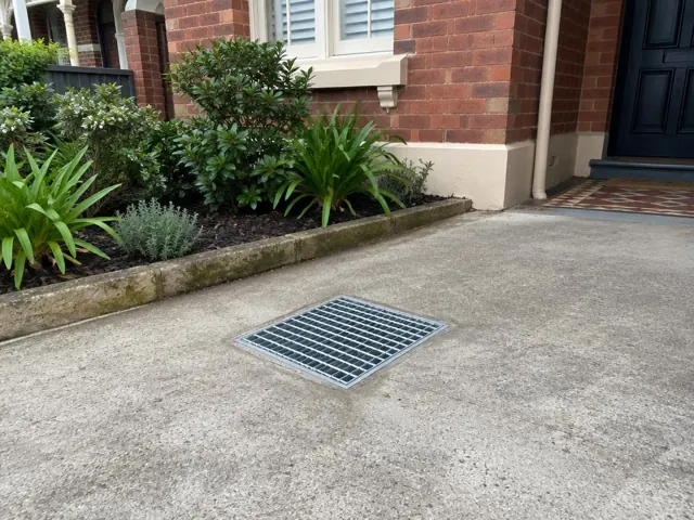 Concrete driveway with a seated stormwater grate beside brick house and garden beds in Kingsford