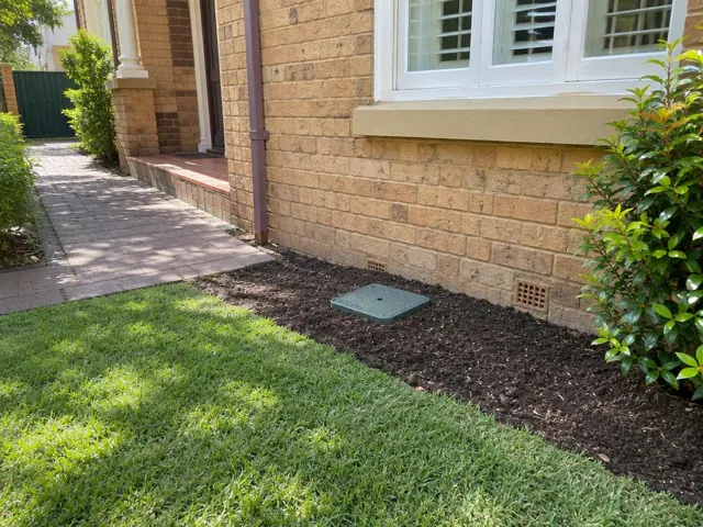 Rectangular green access cover in a landscaped side yard beside a brick house in Kensington